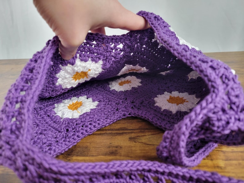 May include: Purple crochet tote bag with white daisy flower accents. The bag is open and the inside is visible.