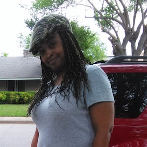 May include: A woman with long, dark hair is wearing a gray shirt and a camouflage-patterned baseball cap. She is standing in front of a red car.