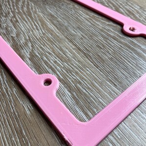 K&ouml;nnte beinhalten: Ein rosafarbener Nummernschildrahmen aus Kunststoff. Der Rahmen hat abgerundete Ecken und eine glatte Oberfl&auml;che.