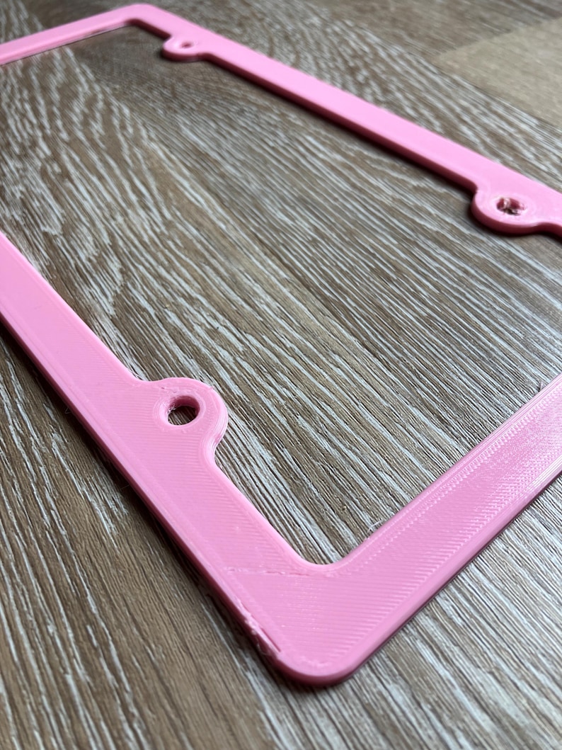 K&ouml;nnte beinhalten: Ein rosafarbener Nummernschildrahmen aus Kunststoff. Der Rahmen hat abgerundete Ecken und eine glatte Oberfl&auml;che.