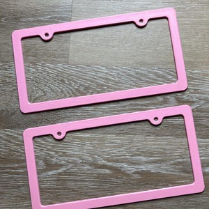 K&ouml;nnte beinhalten: Zwei rosafarbene Nummernschildrahmen aus Kunststoff. Die Rahmen sind rechteckig und haben vier L&ouml;cher f&uuml;r Schrauben.