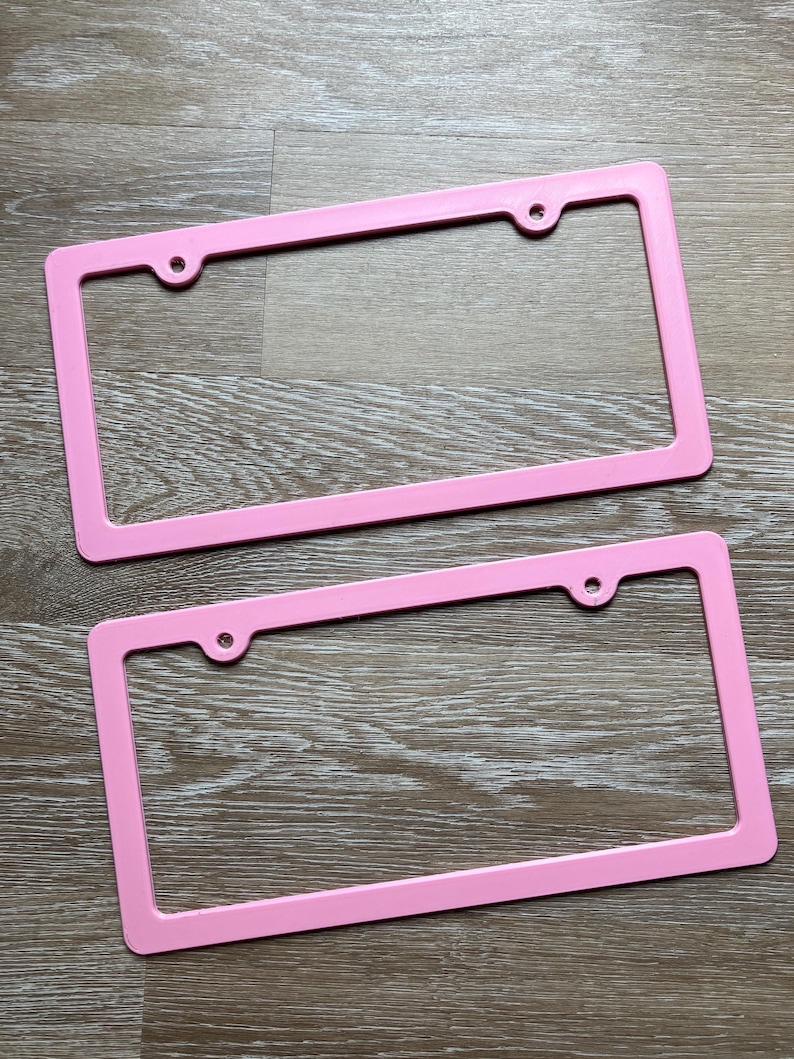 K&ouml;nnte beinhalten: Zwei rosafarbene Nummernschildrahmen aus Kunststoff. Die Rahmen sind rechteckig und haben vier L&ouml;cher f&uuml;r Schrauben.