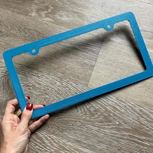 May include: A teal blue plastic license plate frame with rounded corners and two holes on the top.