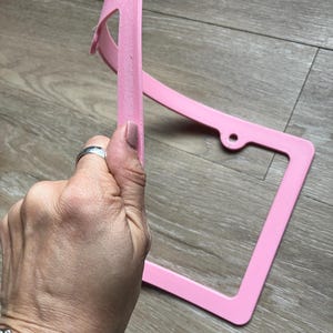 K&ouml;nnte beinhalten: Ein rosafarbener Nummernschildrahmen aus Kunststoff mit abgerundeter Oberkante. Der Rahmen ist leicht verbogen und hat ein kleines Loch auf der rechten Seite.