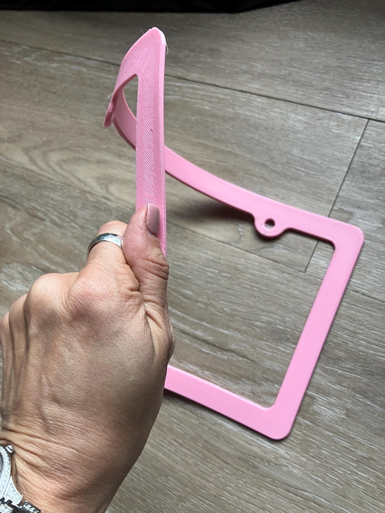 K&ouml;nnte beinhalten: Ein rosafarbener Nummernschildrahmen aus Kunststoff mit abgerundeter Oberkante. Der Rahmen ist leicht verbogen und hat ein kleines Loch auf der rechten Seite.