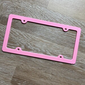 K&ouml;nnte beinhalten: Ein rosafarbener Nummernschildrahmen aus Kunststoff mit vier L&ouml;chern f&uuml;r Befestigungsschrauben.