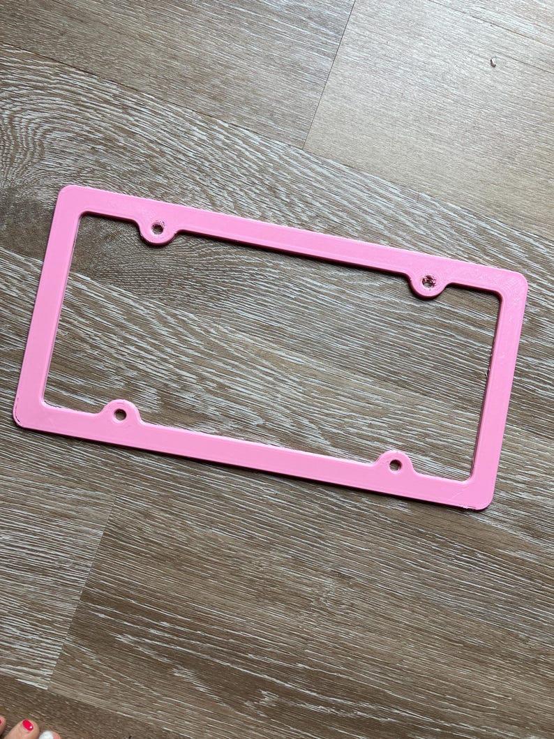 K&ouml;nnte beinhalten: Ein rosafarbener Nummernschildrahmen aus Kunststoff mit vier L&ouml;chern f&uuml;r Befestigungsschrauben.