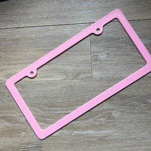 K&ouml;nnte beinhalten: Ein rosafarbener Nummernschildrahmen aus Kunststoff mit zwei L&ouml;chern oben und zwei L&ouml;chern unten.