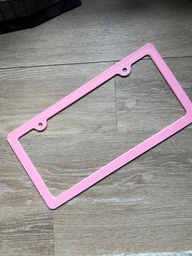 K&ouml;nnte beinhalten: Ein rosafarbener Nummernschildrahmen aus Kunststoff mit zwei L&ouml;chern oben und zwei L&ouml;chern unten.