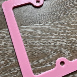 K&ouml;nnte beinhalten: Ein rosafarbener Nummernschildrahmen aus Kunststoff mit zwei L&ouml;chern zur Befestigung. Der Rahmen hat eine strukturierte Oberfl&auml;che.