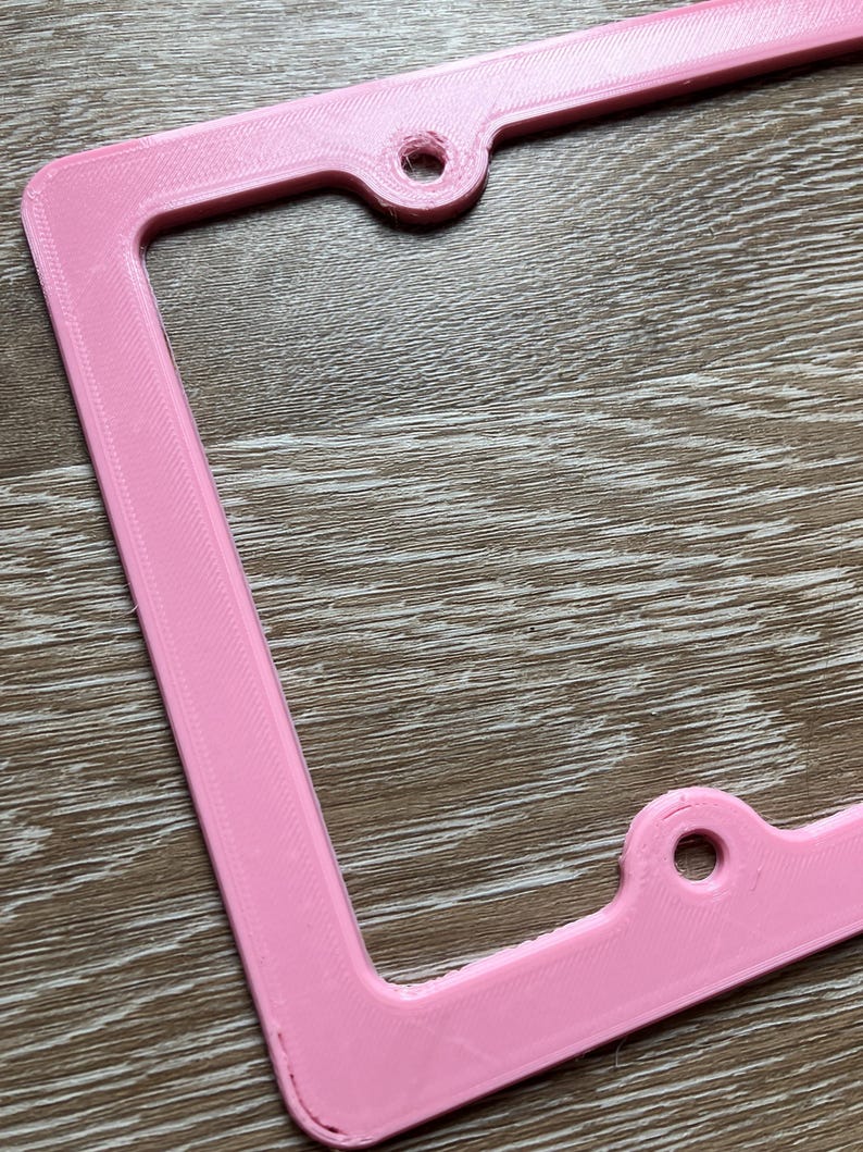 K&ouml;nnte beinhalten: Ein rosafarbener Nummernschildrahmen aus Kunststoff mit zwei L&ouml;chern zur Befestigung. Der Rahmen hat eine strukturierte Oberfl&auml;che.