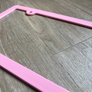 K&ouml;nnte beinhalten: Ein rosafarbener Nummernschildrahmen aus Kunststoff. Der Rahmen besteht aus glattem, gl&auml;nzendem Kunststoff und hat ein schlichtes, modernes Design.