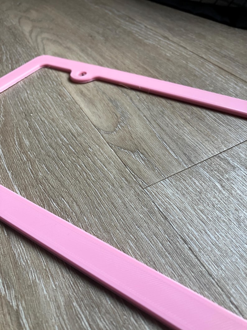 K&ouml;nnte beinhalten: Ein rosafarbener Nummernschildrahmen aus Kunststoff. Der Rahmen besteht aus glattem, gl&auml;nzendem Kunststoff und hat ein schlichtes, modernes Design.