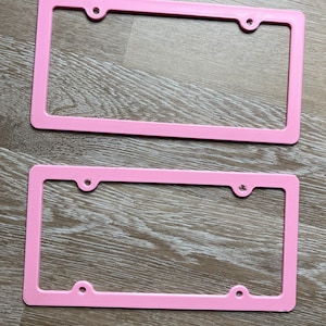 K&ouml;nnte beinhalten: Zwei rosa Kunststoff-Kennzeichenrahmen. Die Rahmen sind rechteckig mit abgerundeten Ecken und haben vier L&ouml;cher f&uuml;r Befestigungsschrauben.