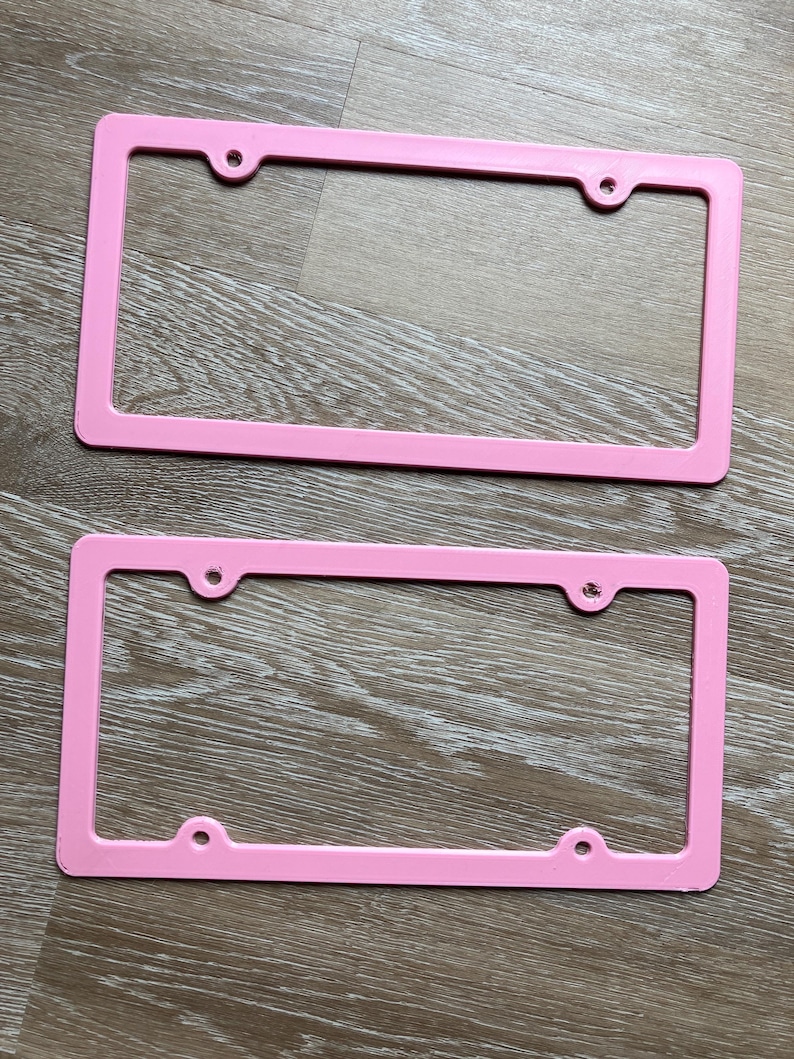 K&ouml;nnte beinhalten: Zwei rosa Kunststoff-Kennzeichenrahmen. Die Rahmen sind rechteckig mit abgerundeten Ecken und haben vier L&ouml;cher f&uuml;r Befestigungsschrauben.