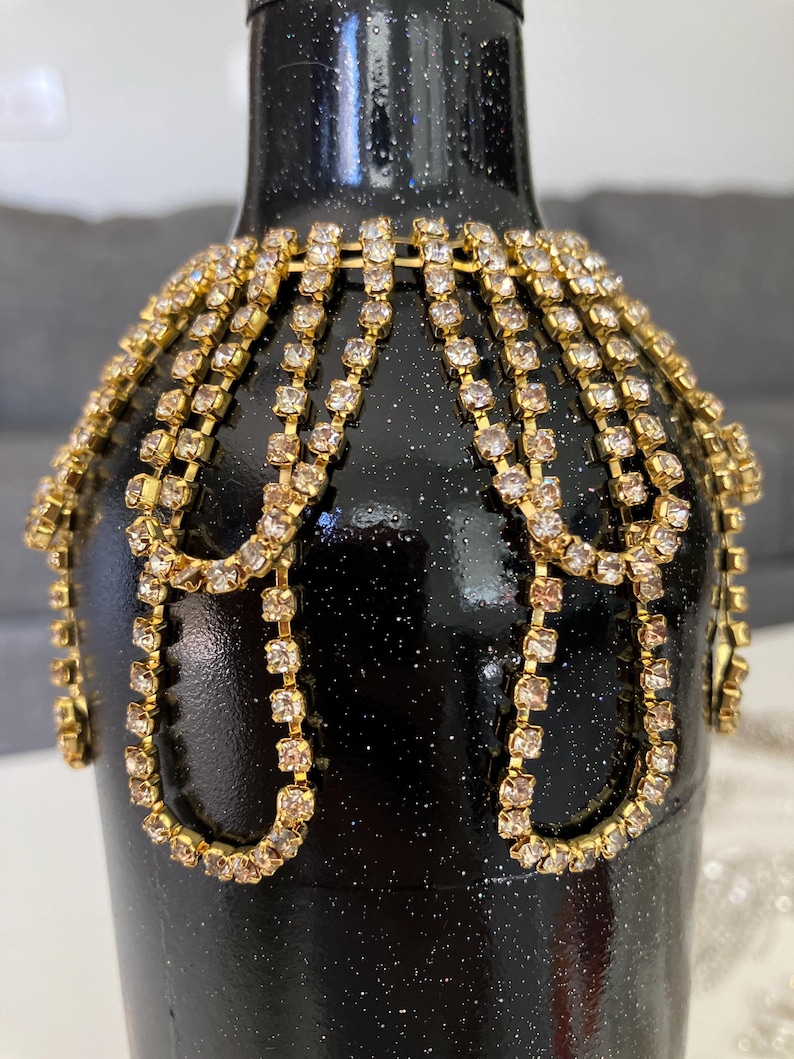May include: A black glass bottle with a sparkly finish, decorated with gold chains of rhinestones. The bottle is a unique and decorative piece.