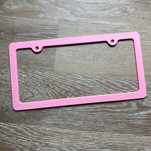 K&ouml;nnte beinhalten: Ein rosafarbener Nummernschildrahmen aus Kunststoff mit zwei L&ouml;chern f&uuml;r Schrauben.