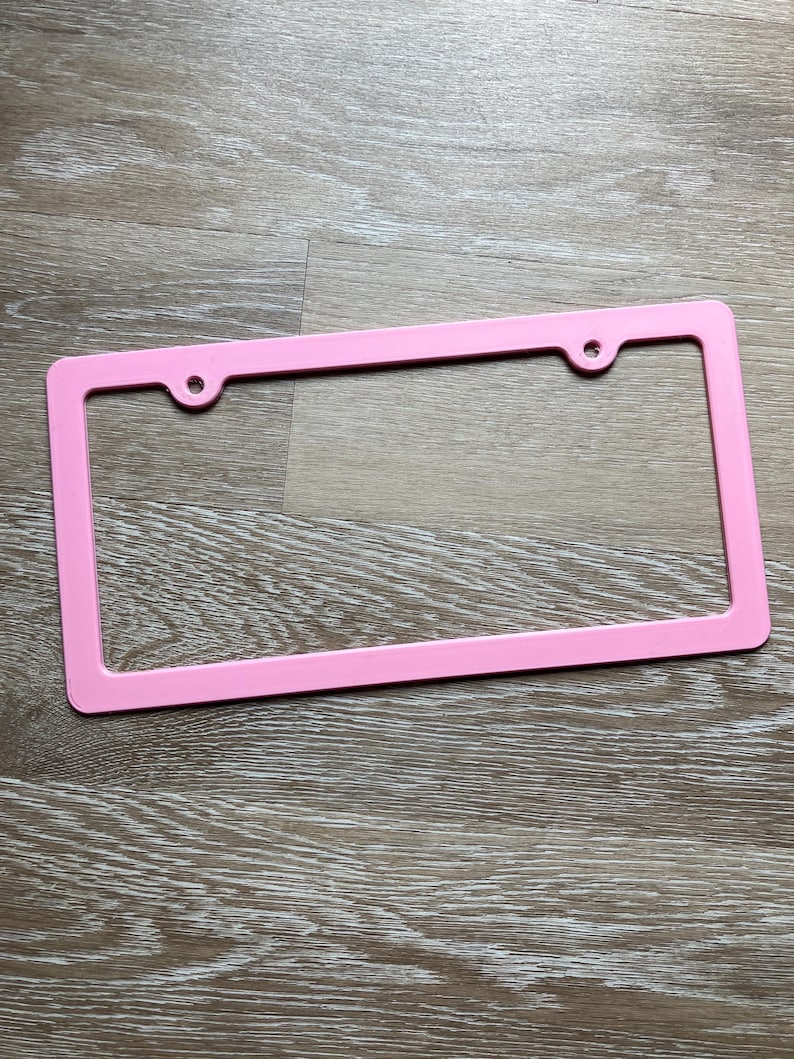 K&ouml;nnte beinhalten: Ein rosafarbener Nummernschildrahmen aus Kunststoff mit zwei L&ouml;chern f&uuml;r Schrauben.