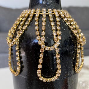 May include: A black glass bottle decorated with gold and clear rhinestone chains. The bottle has a sparkly black finish.