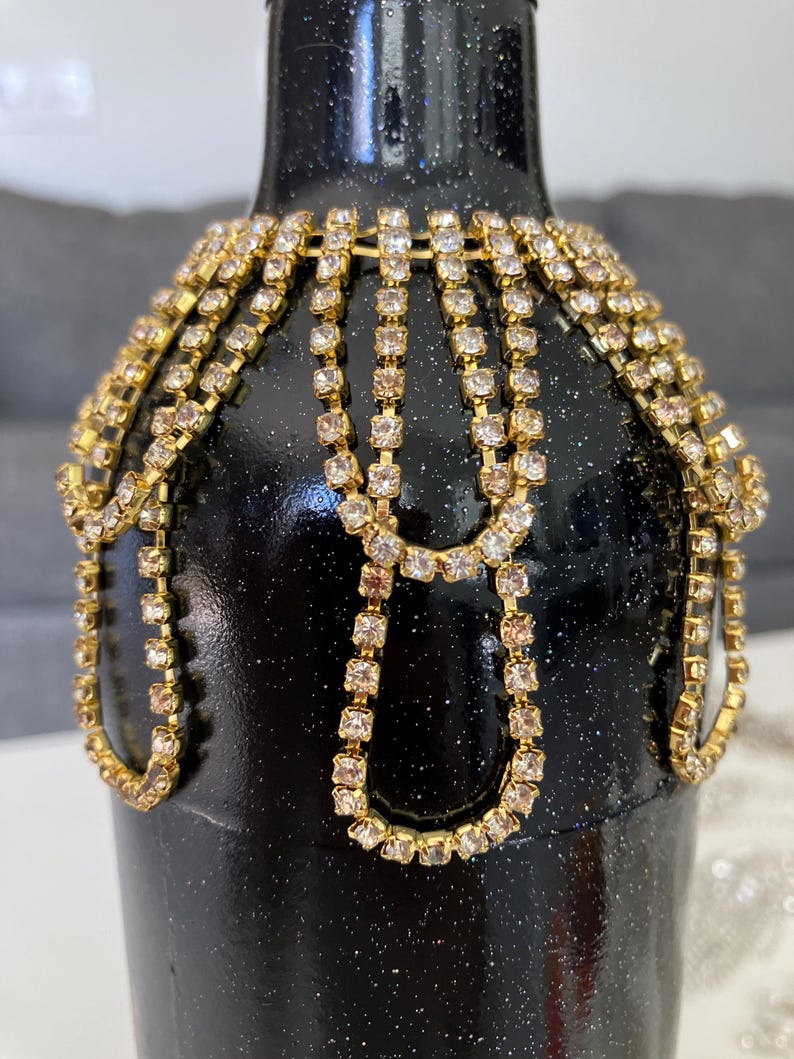 May include: A black glass bottle decorated with gold and clear rhinestone chains. The bottle has a sparkly black finish.