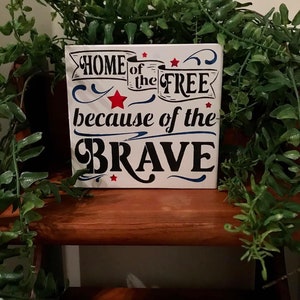 May include: White wooden sign with blue and red accents. The sign reads "Home of the Free because of the Brave".