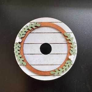 May include: A round, wooden decorative piece with a white, wood-planked background. A brown ring encircles a black hole in the center. Green leaf and white flower cutouts adorn the outer edge of the brown ring, creating a wreath-like design.