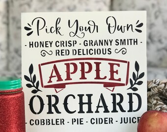 May include: A white wooden sign with black lettering that reads "Pick Your Own Honey Crisp - Granny Smith - Red Delicious Apple Orchard Cobbler - Pie - Cider - Juice". The sign has a red apple graphic with green leaves on either side.