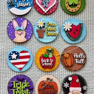 May include: A collection of twelve round wooden decorations with various seasonal themes. Designs include a snowflake, heart, bunny, watermelon, American flag heart, apple, pumpkin, and gnome. Each piece features text like "Freezin' Season" and "Trick or Treat".