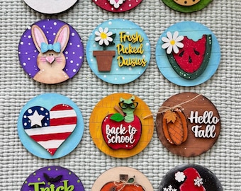 Può includere: Una collezione di dodici decorazioni rotonde in legno con vari temi stagionali. I disegni includono un fiocco di neve, un cuore, un coniglio, un'anguria, un cuore con la bandiera americana, una mela, una zucca e uno gnomo. Con testi come "Freezin' Season".
