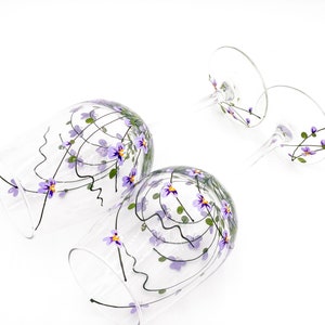 K&ouml;nnte beinhalten: Zwei klare Weingl&auml;ser aus Glas mit auf die Oberfl&auml;che gemalten violetten und gr&uuml;nen Blumenmustern. Die Gl&auml;ser sind leer und haben einen Stiel und einen runden Boden.