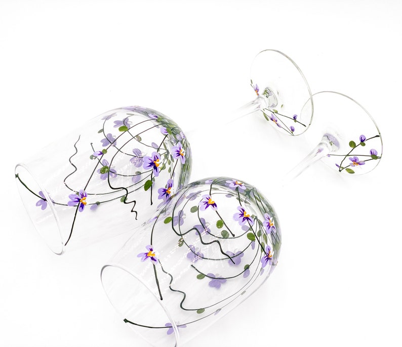 K&ouml;nnte beinhalten: Zwei klare Weingl&auml;ser aus Glas mit auf die Oberfl&auml;che gemalten violetten und gr&uuml;nen Blumenmustern. Die Gl&auml;ser sind leer und haben einen Stiel und einen runden Boden.