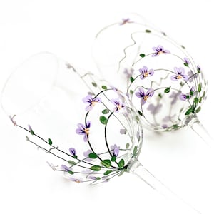 K&ouml;nnte beinhalten: Zwei klare Weingl&auml;ser aus Glas mit handbemalten lila Bl&uuml;ten und gr&uuml;nen Bl&auml;ttern.