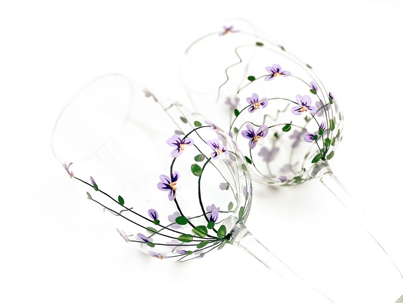 K&ouml;nnte beinhalten: Zwei klare Weingl&auml;ser aus Glas mit handbemalten lila Bl&uuml;ten und gr&uuml;nen Bl&auml;ttern.