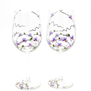 K&ouml;nnte beinhalten: Zwei klare Weingl&auml;ser aus Glas mit handbemaltem Design aus lila Blumen und Ranken. Die Blumen sind helllila mit gelben Zentren.