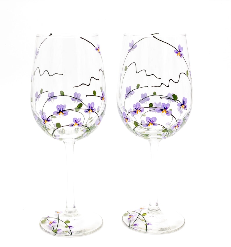 K&ouml;nnte beinhalten: Zwei klare Weingl&auml;ser aus Glas mit handbemaltem Design aus lila Blumen und Ranken. Die Blumen sind helllila mit gelben Zentren.