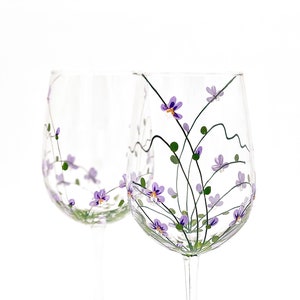 K&ouml;nnte beinhalten: Zwei klare Weingl&auml;ser aus Glas mit handbemalten lila Bl&uuml;ten und gr&uuml;nen Stielen. Die Bl&uuml;ten sind in einem zarten Muster zusammengef&uuml;gt.