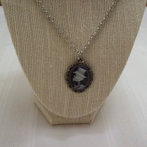 May include: A silver chain necklace with a cameo pendant featuring a black and white image of a person wearing a top hat. The cameo is set in a silver frame with a decorative border.