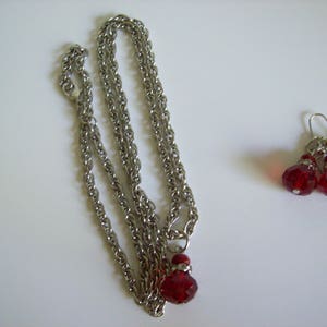 Può includere: Una collana color argento con un pendente rosso e un paio di orecchini abbinati. La collana ha un design a catena e il pendente e gli orecchini presentano perline rosse sfaccettate. Il set di gioielli è esposto su una superficie bianca.
