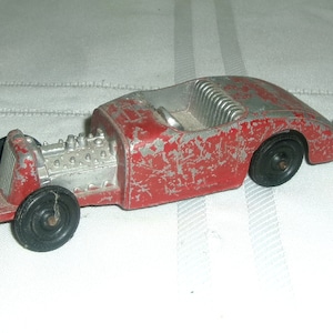 May include: A red and silver toy car with a visible engine and black rubber wheels. The car has a worn, vintage look.