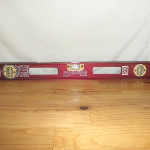 May include: A red level tool with gold accents and the text "Sheffield Keen England". The level has two circular bubble vials and a rectangular vial in the center. The tool is resting on a wooden surface.