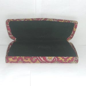 May include: A red, yellow, and gold patterned fabric eyeglasses case with a black velvet interior. The case is open and the interior is visible.