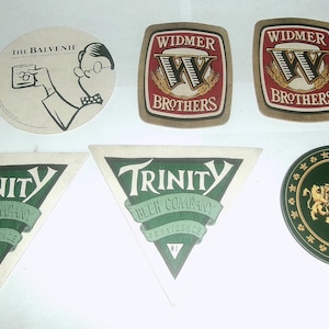 May include: A collection of ten coasters featuring various brewery logos. The coasters are round and square, with a variety of colors and designs. The coasters feature the names "The Balvenie", "Widmer Brothers", and "Trinity Beer Company".