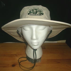 May include: A beige sun hat with a wide brim, featuring the text "Overseas Adventure Travel Sir Edmund Hillary Club" in green. The hat has a black chin strap and is displayed on a white mannequin head.