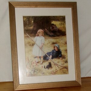 May include: Framed print of a painting depicting two girls in a field with a black cat. The girl on the left is holding a rake and the girl on the right is lying down. The painting is set in a rural landscape with trees and a grassy field.