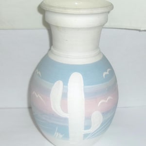 May include: A white ceramic vase with a blue and pink desert scene and a white cactus design.