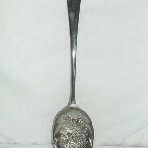 May include: A silver serving spoon with an ornate design featuring a cluster of grapes and leaves. The spoon has a long, slender handle and a rounded bowl.
