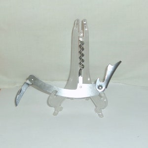 May include: A silver corkscrew with a built-in bottle opener, displayed on a clear plastic stand.