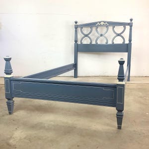 May include: A blue painted bed frame with a headboard. The headboard features an arched design with decorative cutouts. The bed frame has a rectangular shape with decorative trim and four posts. The bed frame is painted blue with white accents.