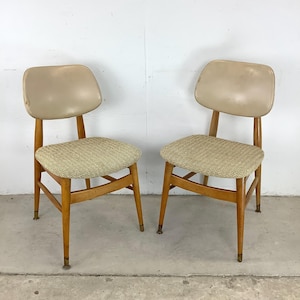 May include: Two mid-century modern chairs. Each chair has a light tan vinyl backrest and a woven seat. The wooden frames are a light brown color, with brass-colored caps on the legs. The chairs are set against a white wall.
