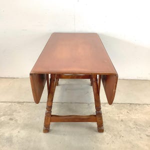 May include: A wooden drop-leaf table with a rectangular top and turned legs. The table is a warm brown color and has two drop leaves that can be raised or lowered. The table is approximately 48 inches long when fully extended.